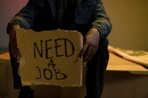 Muž sediaci so sklonenou hlavou a držiaci kartónovú tabuľu s nápisom "Need a Job".