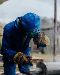 Zvárač v modrom pracovnom odeve a ochrannej prilbe pracujúci so zváračkou v exteriéri.